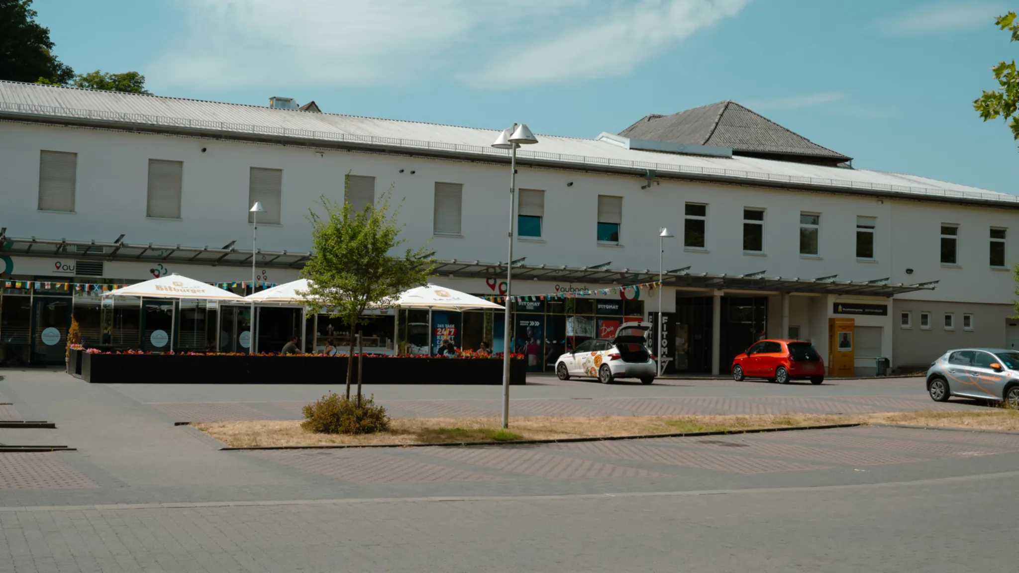 Ladenzeile am Treffpunkt Aubach mit Außenterrasse, Sonnenschirmen und Parkplatz